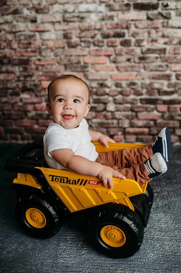Milestone Photography session. A baby boy is sitting in a miniature dump truck and smiling at the camera