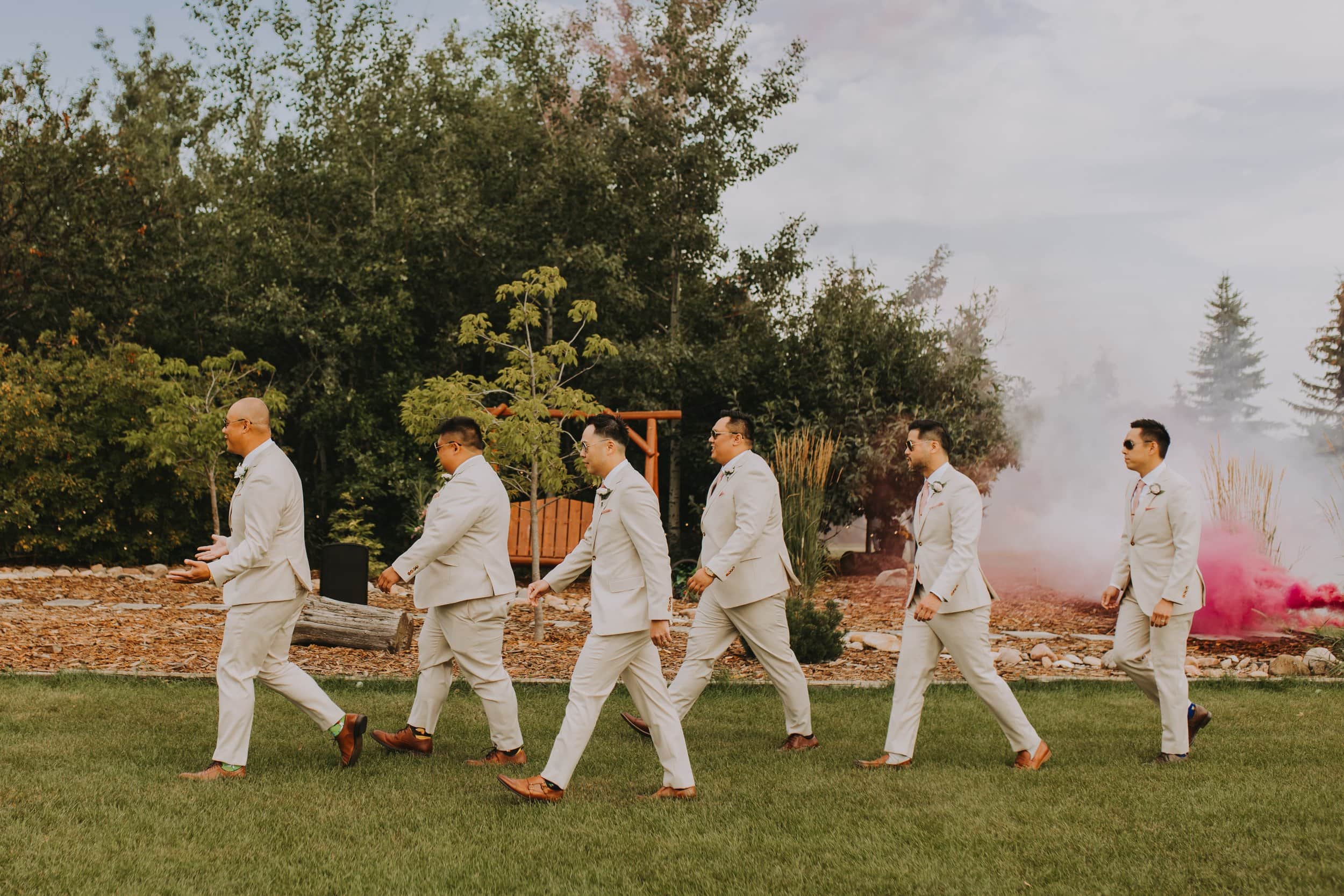 Groomsmen wearing sunglasses and white suits are walking across grass, there is a pink smoke bomb behind them
